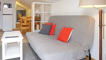 Een moderne woonkamer met een grijze bank, een witte bijzettafel, een lamp en een witte boekenkast met diverse spullen.