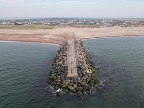 A coastal pier extending into the sea with a sandy beach and houses in the background. - 7 Person Holiday Home in Vestervig (Vestervig)