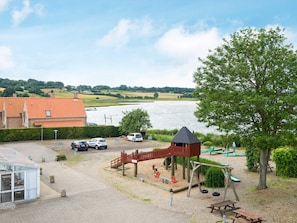 A playground with swings and a climbing structure near a body of water, with a parking area and buildings in the background.