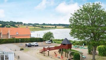 A playground with swings and a climbing structure, a parking area with cars, and a view of a lake.