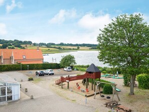 A playground with swings and a climbing structure, a parking area with cars, and a view of a lake. - 4 Person Holiday Home in Hejls (Hejls)