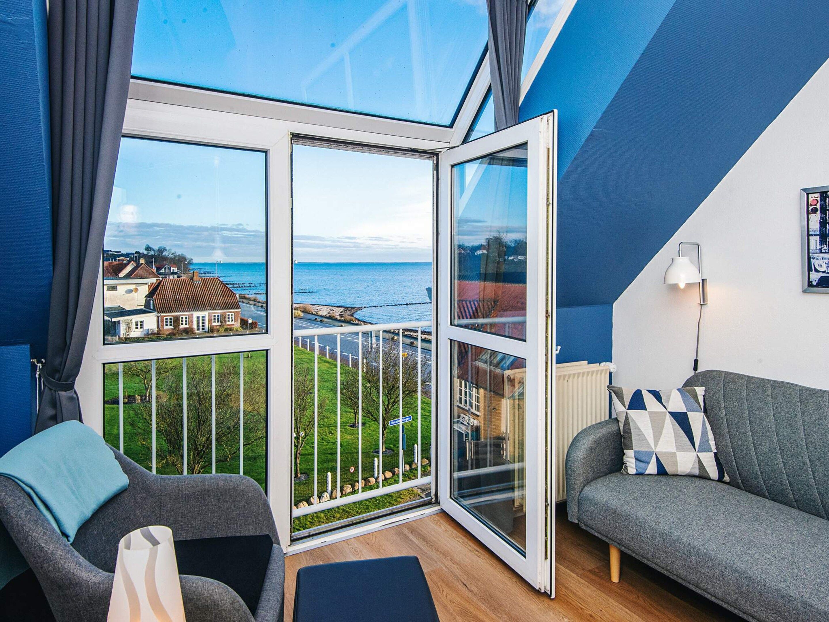 A room with a large sliding glass door leading to a balcony with a view of the sea and buildings. There is a grey sofa and a chair with a cushion.