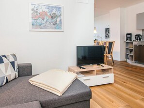 Una sala de estar moderna con un sofá gris, un televisor de pantalla plana sobre un soporte de madera, una zona de comedor con una mesa y sillas de madera, y una cocina con una cafetera y un lavavajillas.