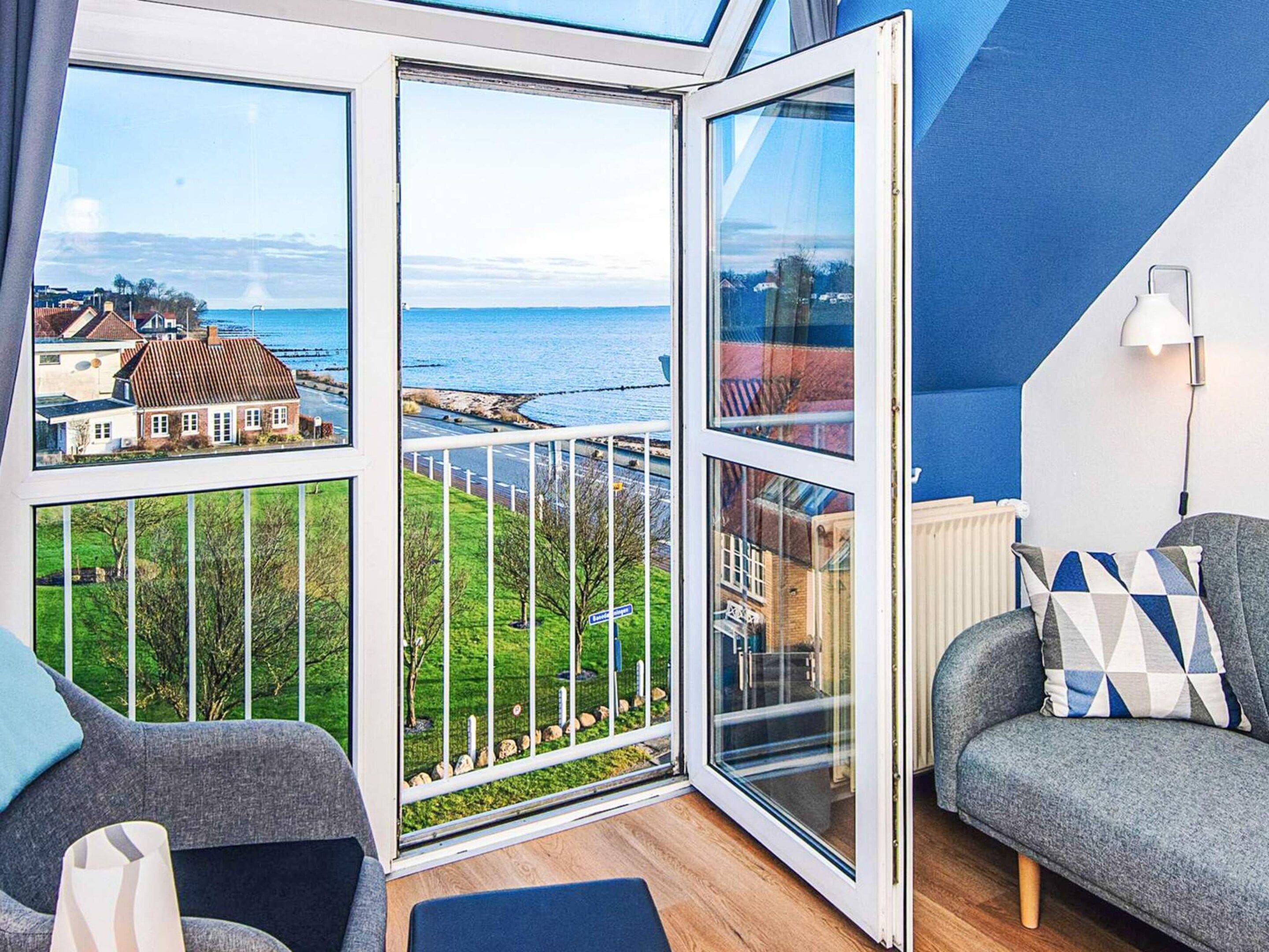 Una habitación con vista al mar, un balcón con barandilla y un sofá gris con cojines estampados.