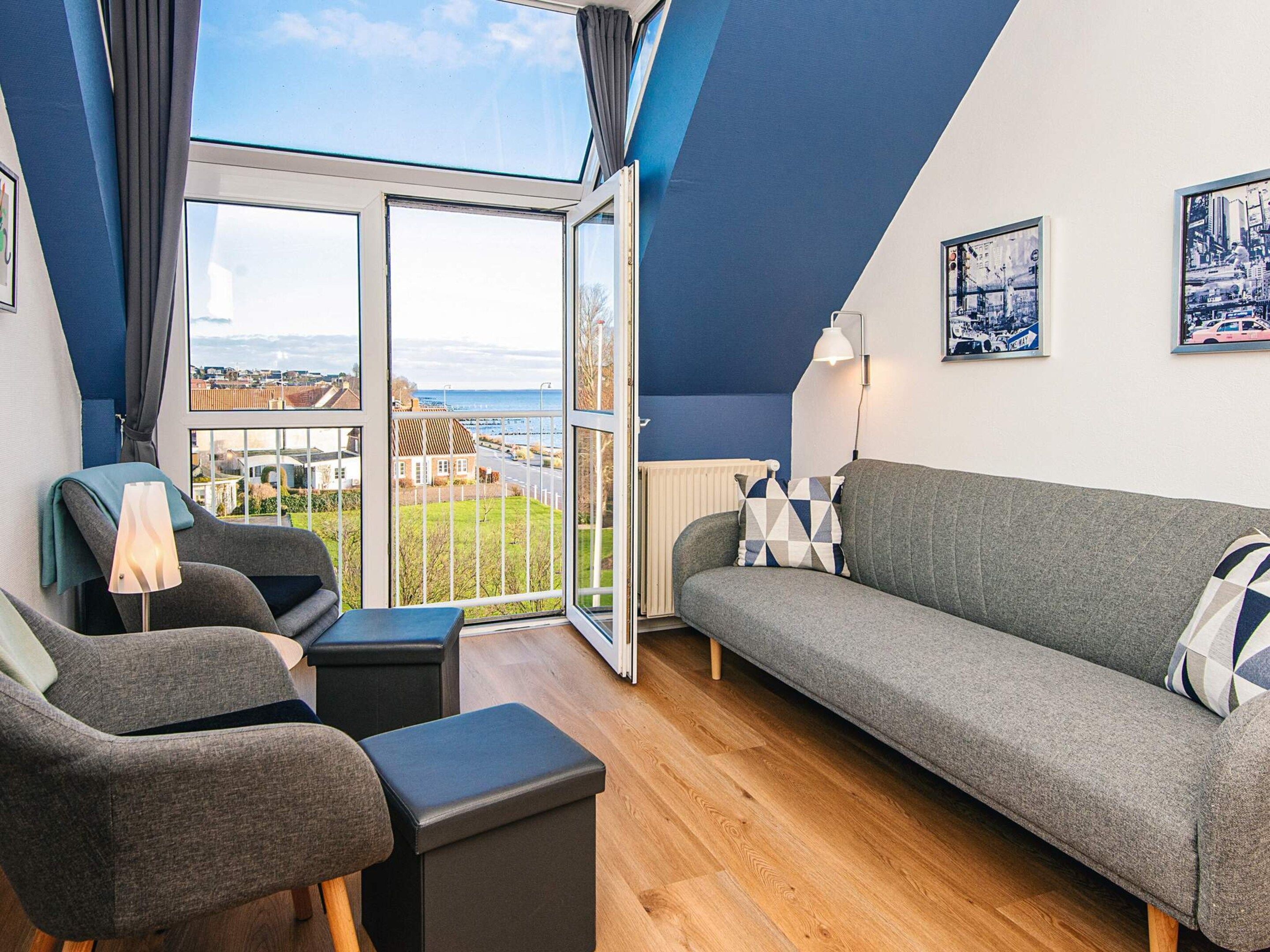A modern living room with a grey sofa, a grey armchair, and a black ottoman. There is a large window with a view of the sea and buildings.