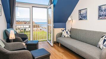 A modern living room with a grey sofa, a grey armchair, and a black ottoman. There is a large window with a view of the sea and buildings.