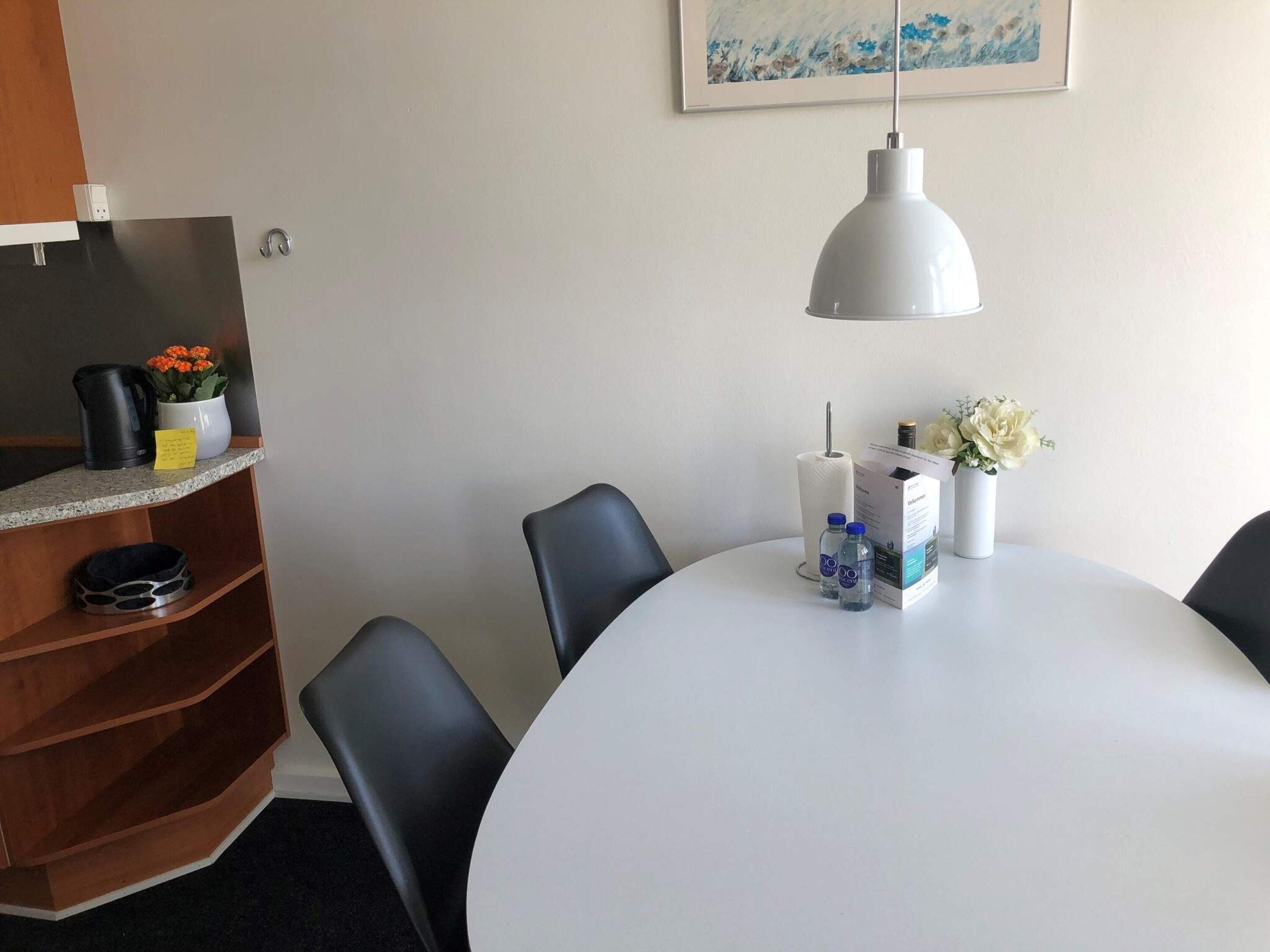 A hotel room with a round table, black chairs, a white lamp, a shelf with a coffee maker, a vase with flowers, and a framed picture on the wall.