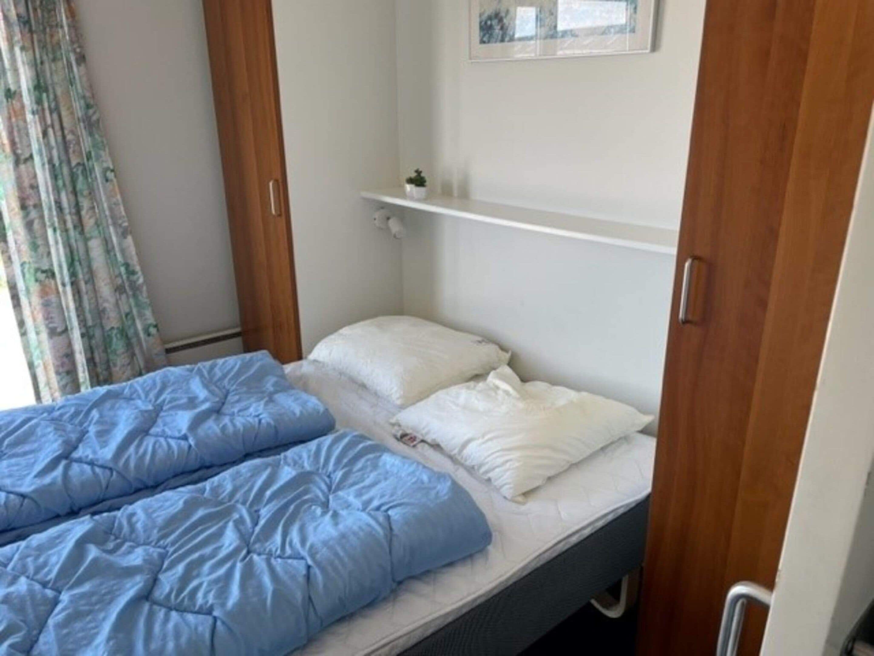 Ein Schlafzimmer mit einem Bett, einer blauen Decke, weißen Kissen, einer Holztür und einem kleinen Regal mit einer Pflanze.