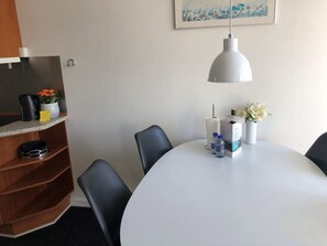 Quarto de hotel com uma mesa redonda, cadeiras pretas, toalha de mesa branca, luminária pendente branca, prateleira com cafeteira, um vaso com flores e um quadro na parede.