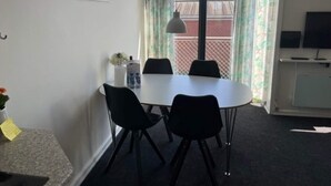 A room with a white table, four black chairs, a small white cabinet, a television, and a window with curtains.