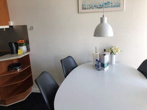 A hotel room with a round table, black chairs, a white lamp, a shelf with a coffee maker, a flower vase, and a painting on the wall.