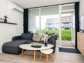 Un soggiorno moderno con un divano grigio, un tavolo bianco con una ciotola decorativa e vista su un edificio attraverso una porta vetrata.