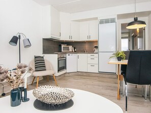 A modern kitchen with white cabinets, a microwave, an oven, and a refrigerator. There is a dining area with a table, chairs, and a potted plant.