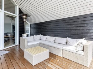 Un espace extérieur aménagé, abrité, avec un canapé en osier blanc et un pouf assorti, un radiateur métallique et une porte en verre donnant accès à un espace intérieur.