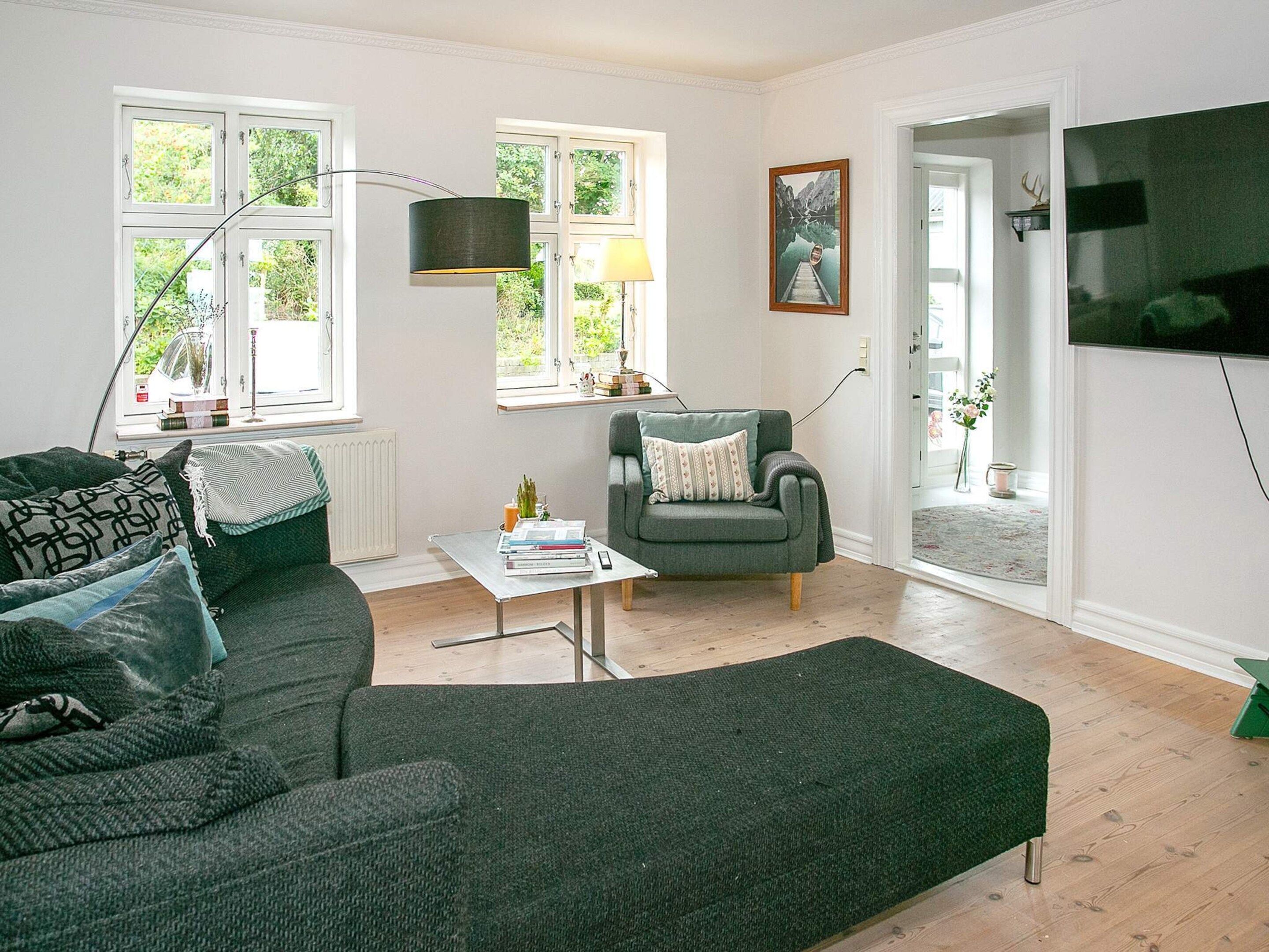 Ein Wohnzimmer mit einer Couch, einem Sessel und einem Couchtisch. Es gibt Fenster, eine Lampe und einen Fernseher.