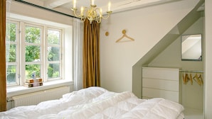 A bedroom with a large bed, a window with curtains, a chandelier, a mirror, and a radiator. - 4 Star Holiday Home in Asperup (Middelfart)