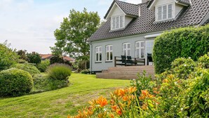 A two-story house with a sloped roof, a front porch with chairs, and a well-maintained garden with various plants and flowers. - 4 Star Holiday Home in Asperup (Middelfart)