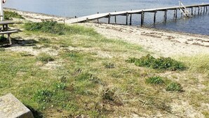 Una playa con un muelle de madera que se adentra en aguas tranquilas, una mesa de picnic y zonas verdes.