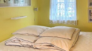 Una cama con dos almohadillas y un cabecero blanco en una habitación con paredes amarillas y una ventana con cortinas de encaje.