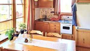 Een keuken met houten kastjes, een fornuis en een spoelbak. Op de vensterbank staan planten in potten en er is een tafel met stoelen.
