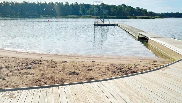 A wooden dock extending into a calm lake with a sandy beach in the foreground and a forested hill in the background. - 6 Person Holiday Home in Monsteras (Monsteras)