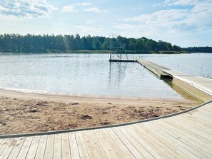 En träbrygga som sträcker sig ut i en stilla sjö, med en sandstrand i förgrunden och en skogbeklädd kulle i bakgrunden.