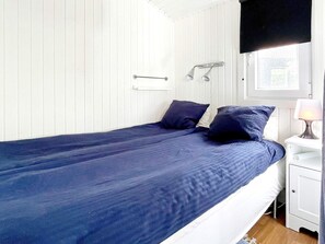 A single bed with a blue bedspread and pillows, a bedside table with a lamp, and a window with a view of greenery outside. - 6 Person Holiday Home in Monsteras (Monsteras)