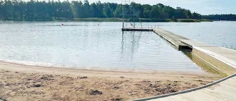 A wooden dock extending into a calm lake with a sandy beach and a forested hill in the background.