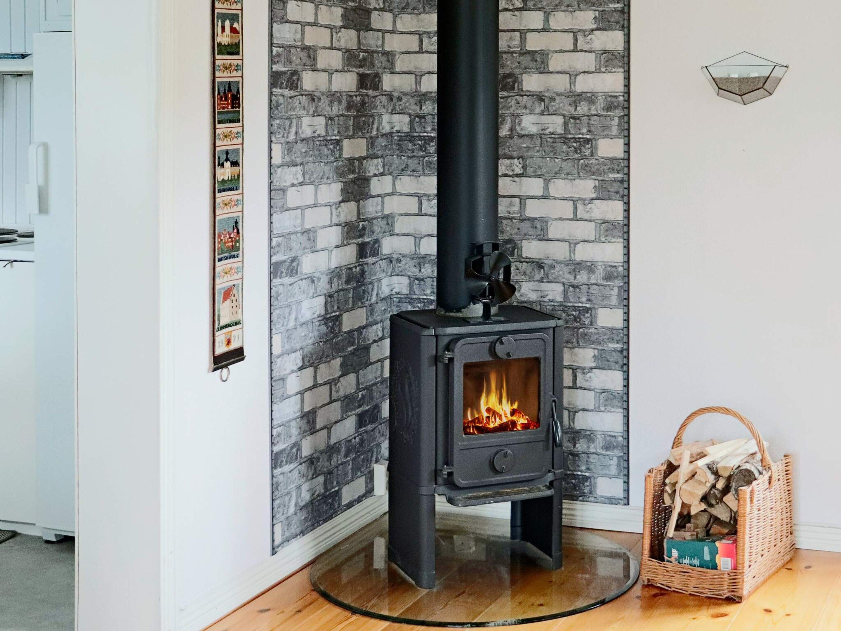 A room with a brick wall, a black wood-burning stove with a glass door, and a wicker basket filled with firewood.