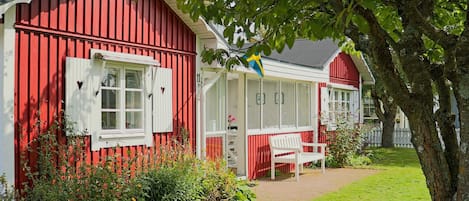 A red wooden house with white shutters, a bench, and a garden area.