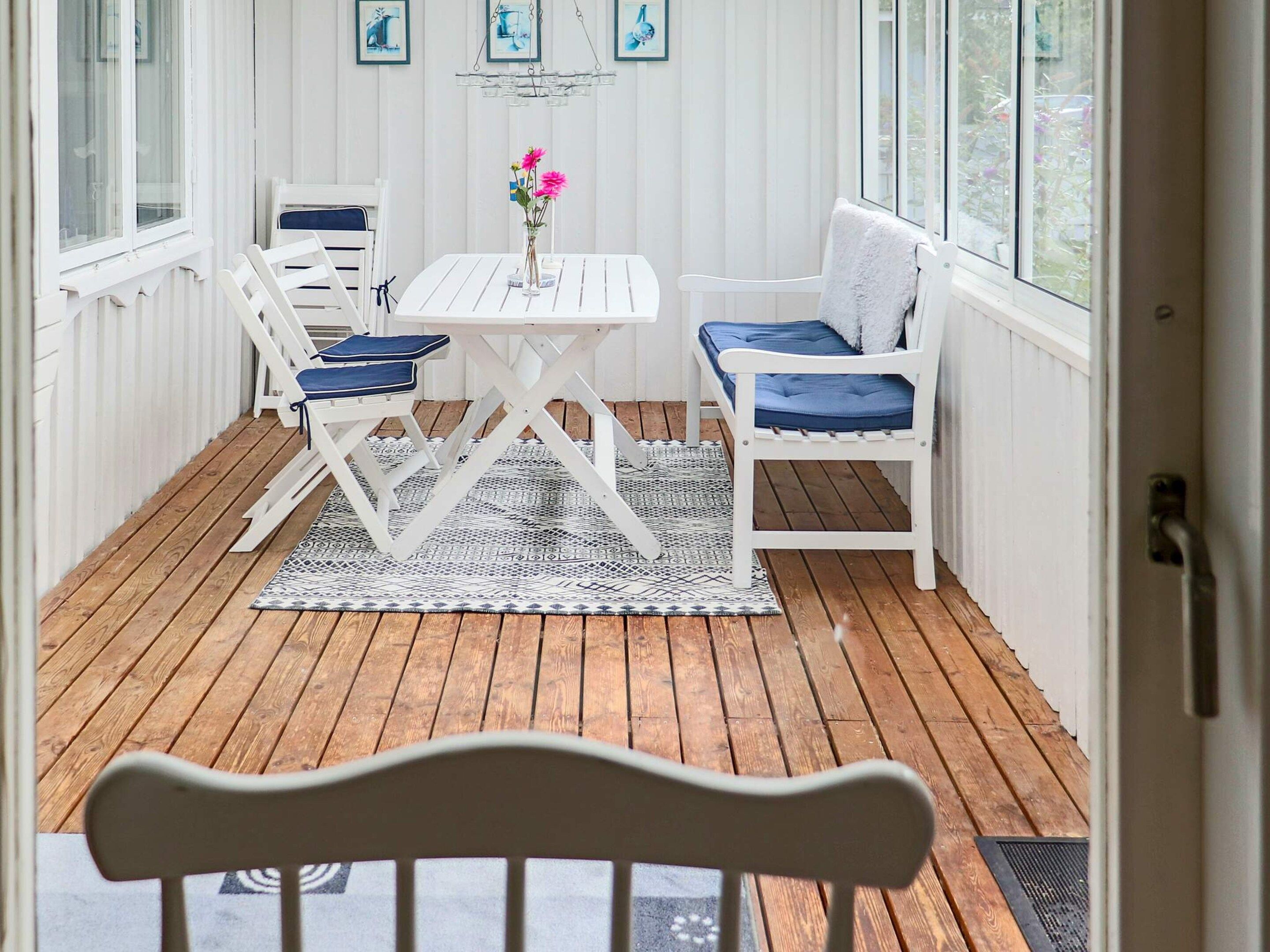 A covered patio with a white table and chairs, a patterned rug, and a view of the outdoors through large windows.