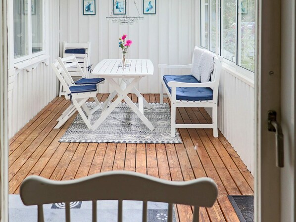 A covered patio with a white table and chairs, a patterned rug, and a view of the outdoors through large windows.
