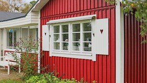 En rød bygning med et hvidt vindue og skodder, omgivet af grønne planter og en bænk.