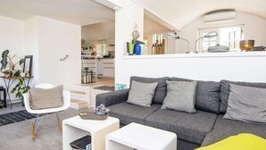 A modern living room with a grey sofa, white side tables, and a yellow throw pillow. There is a white chair with a wicker backrest and a small plant on a side table. A kitchen area is visible in the background. - 4 Star Holiday Home in Grenaa (Grenaa)
