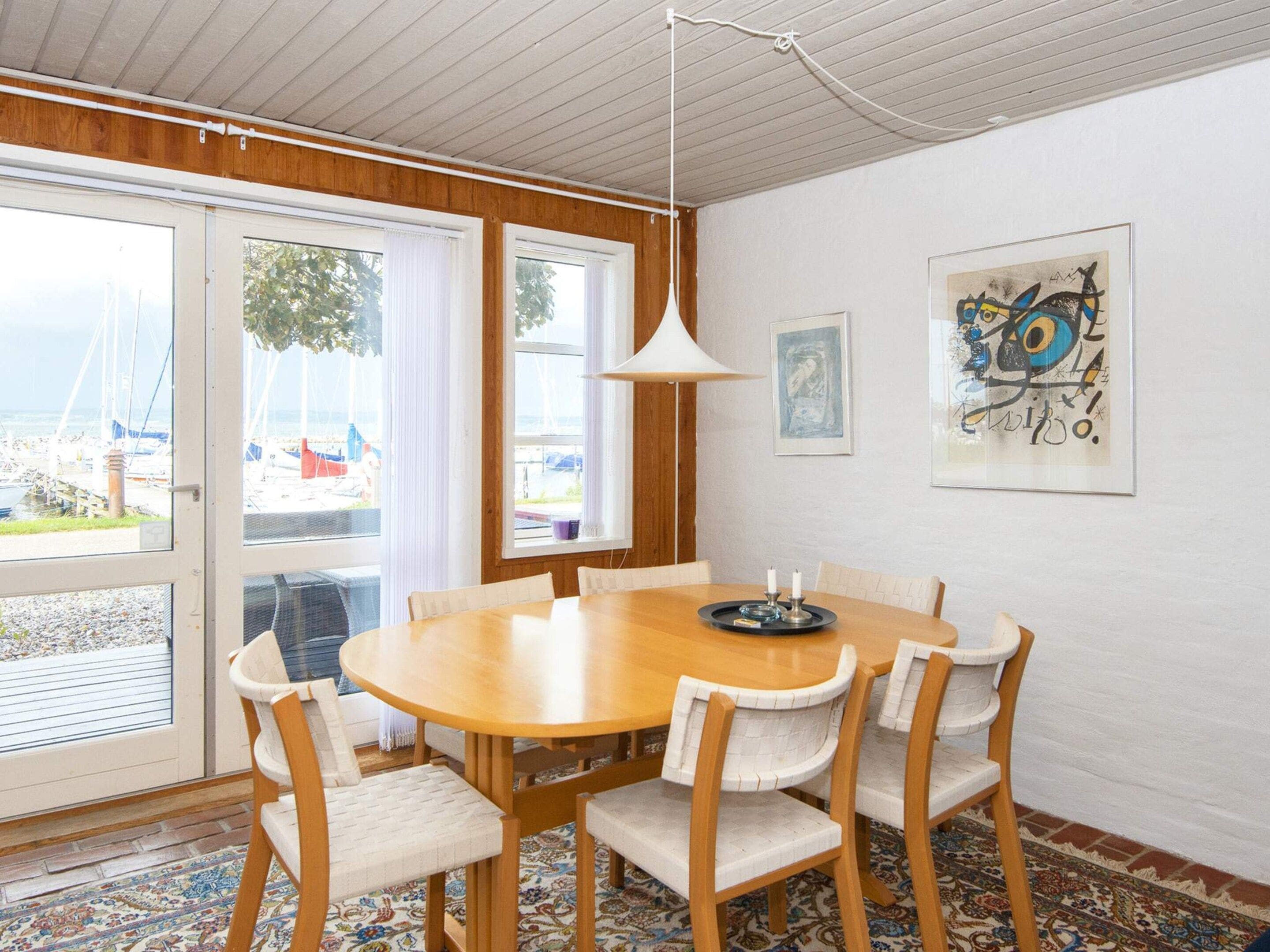 Un comedor con una mesa de madera redonda, seis sillas, una lámpara colgante y vistas amplias al puerto deportivo a través de grandes ventanales.