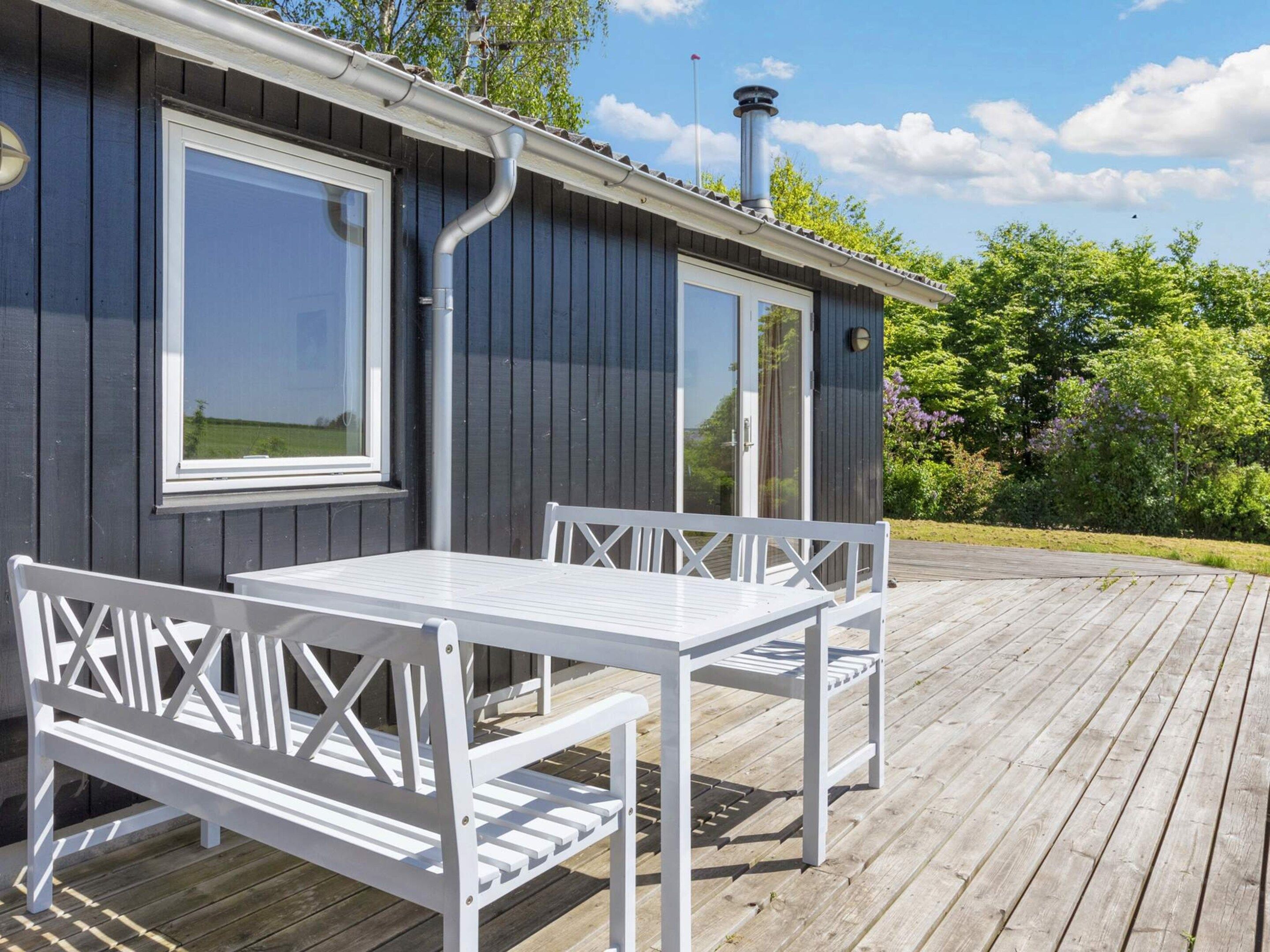 Een houten terras met een witte tafel en bank, een raam en een schoorsteen.