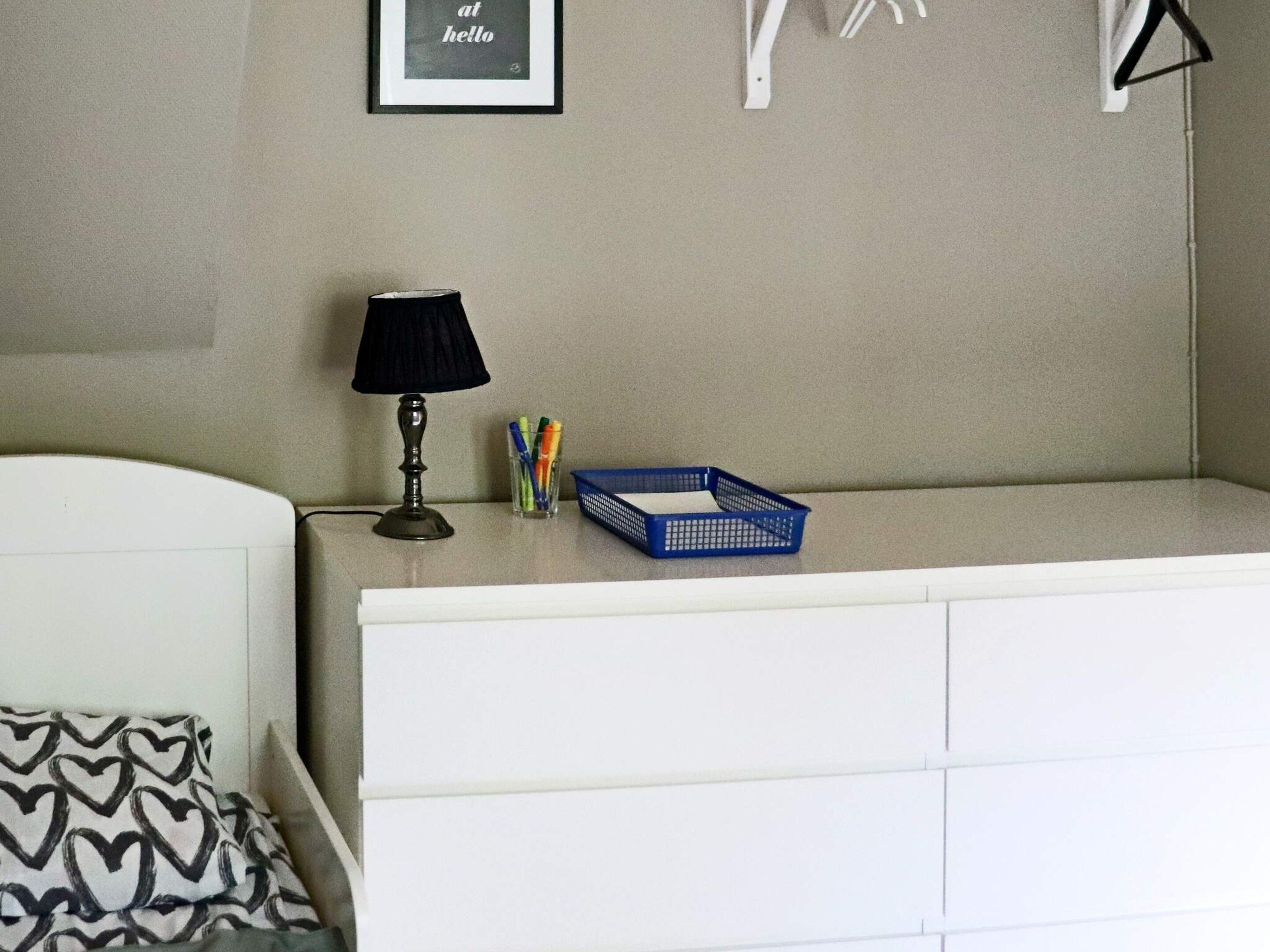 A neatly organized room with a bed, a bedside table with a lamp, a framed picture on the wall, and a blue basket on the dresser.