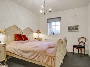 Un dormitorio con cama, mesita de noche, silla, ventana y candelabro.