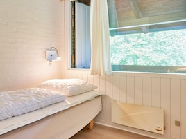 Una habitación pequeña con cama individual, techo inclinado, ventana con cortinas, lámpara de pared y radiador.