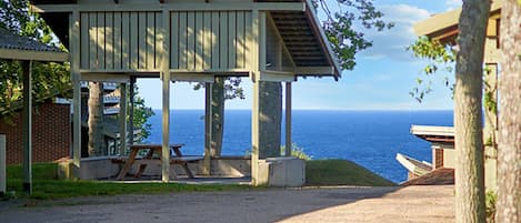 A covered pavilion with a view of the ocean, picnic tables, and a brick building in the background.