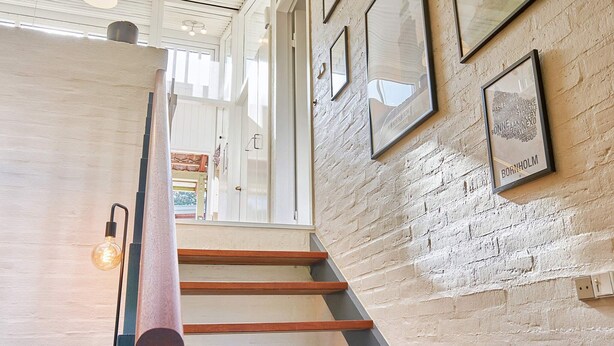 Un escalier avec des marches en bois et un luminaire sur le côté gauche, menant à un niveau supérieur doté d’un plafond blanc et de tableaux encadrés accrochés aux murs.