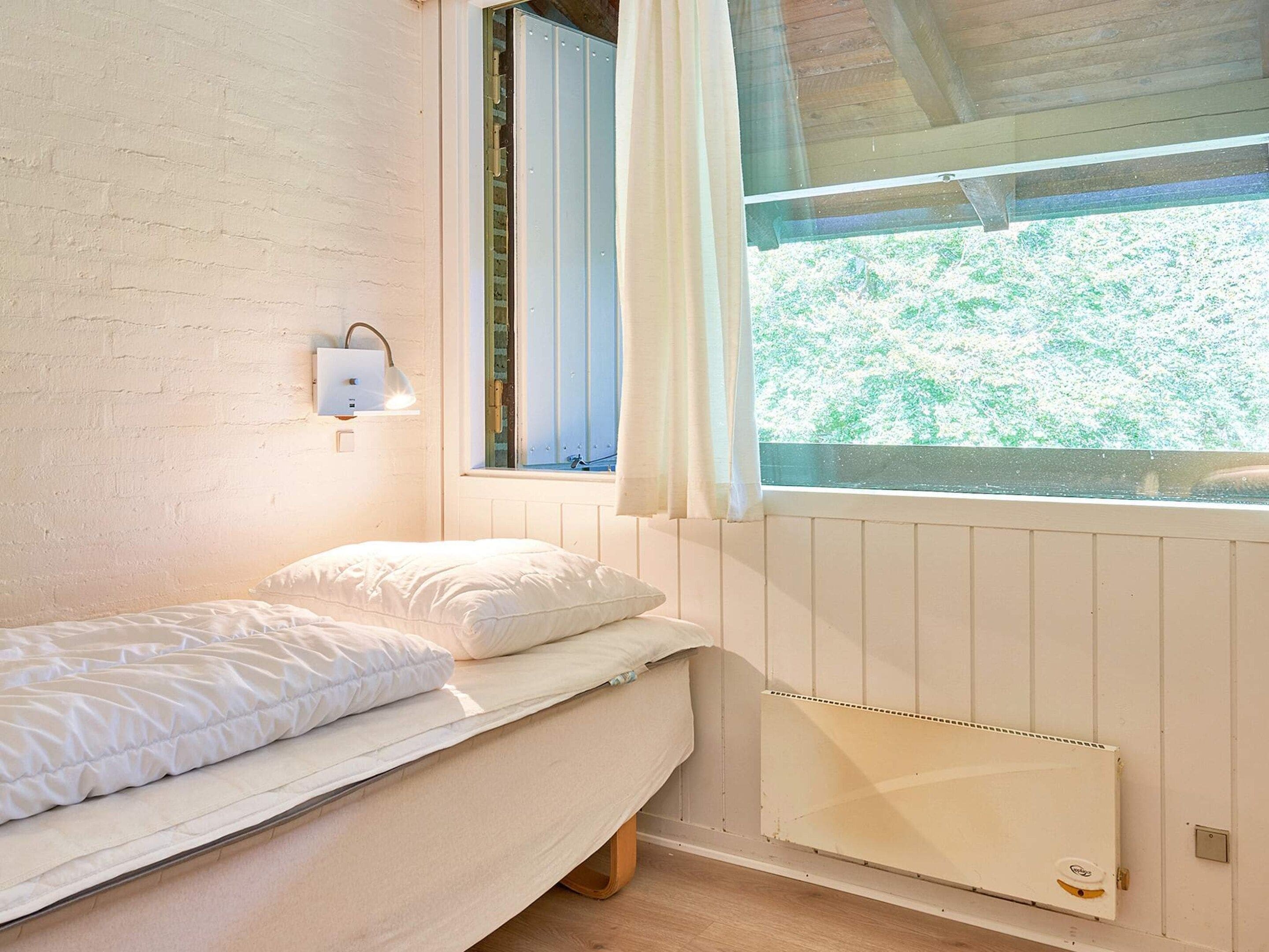 A small, single-bed room with a wooden floor, a white radiator, a window with curtains, and a wall-mounted light fixture.