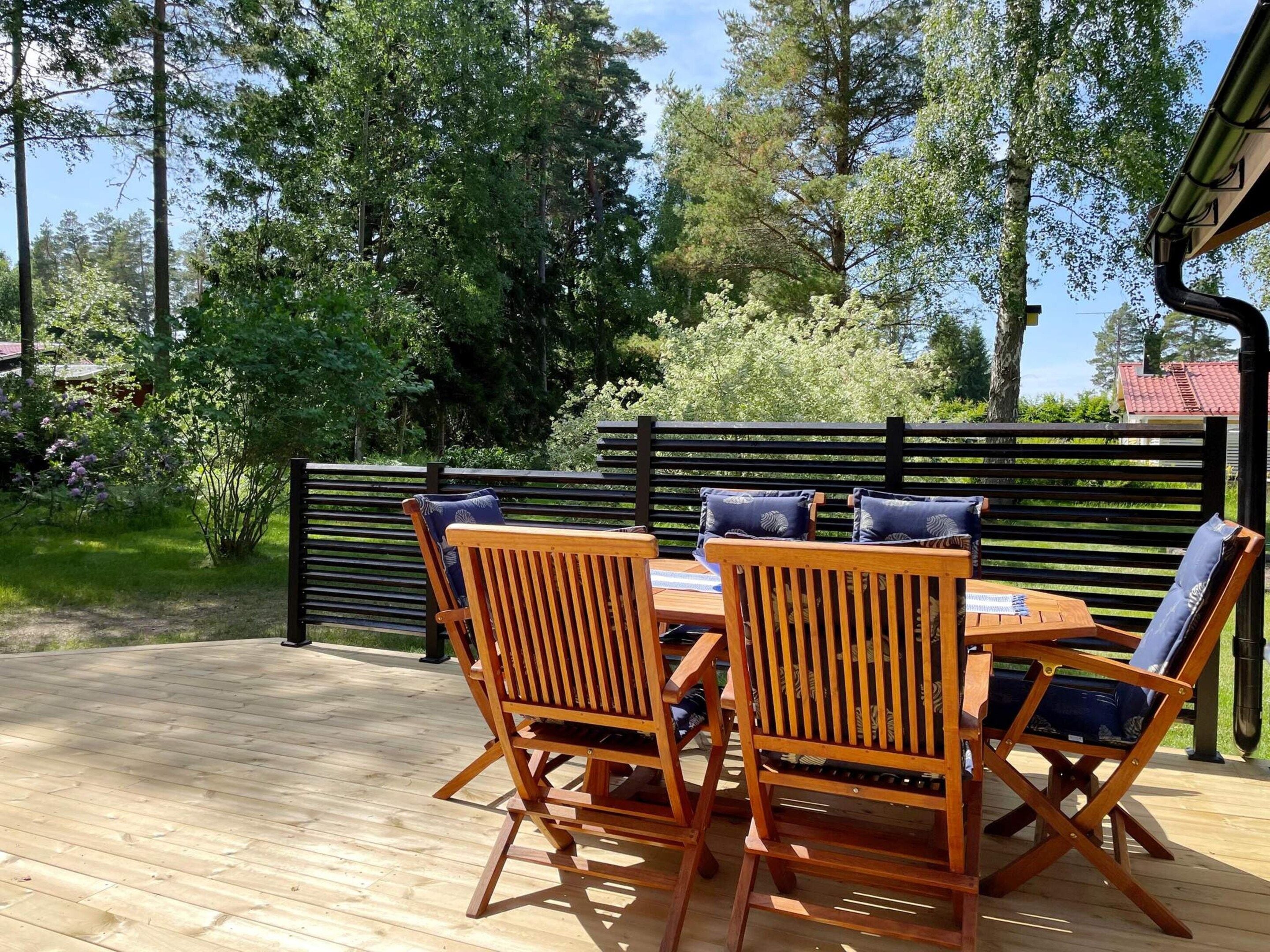 Una terraza de madera al aire libre con una mesa y cuatro sillas, rodeada por una cerca negra y árboles.