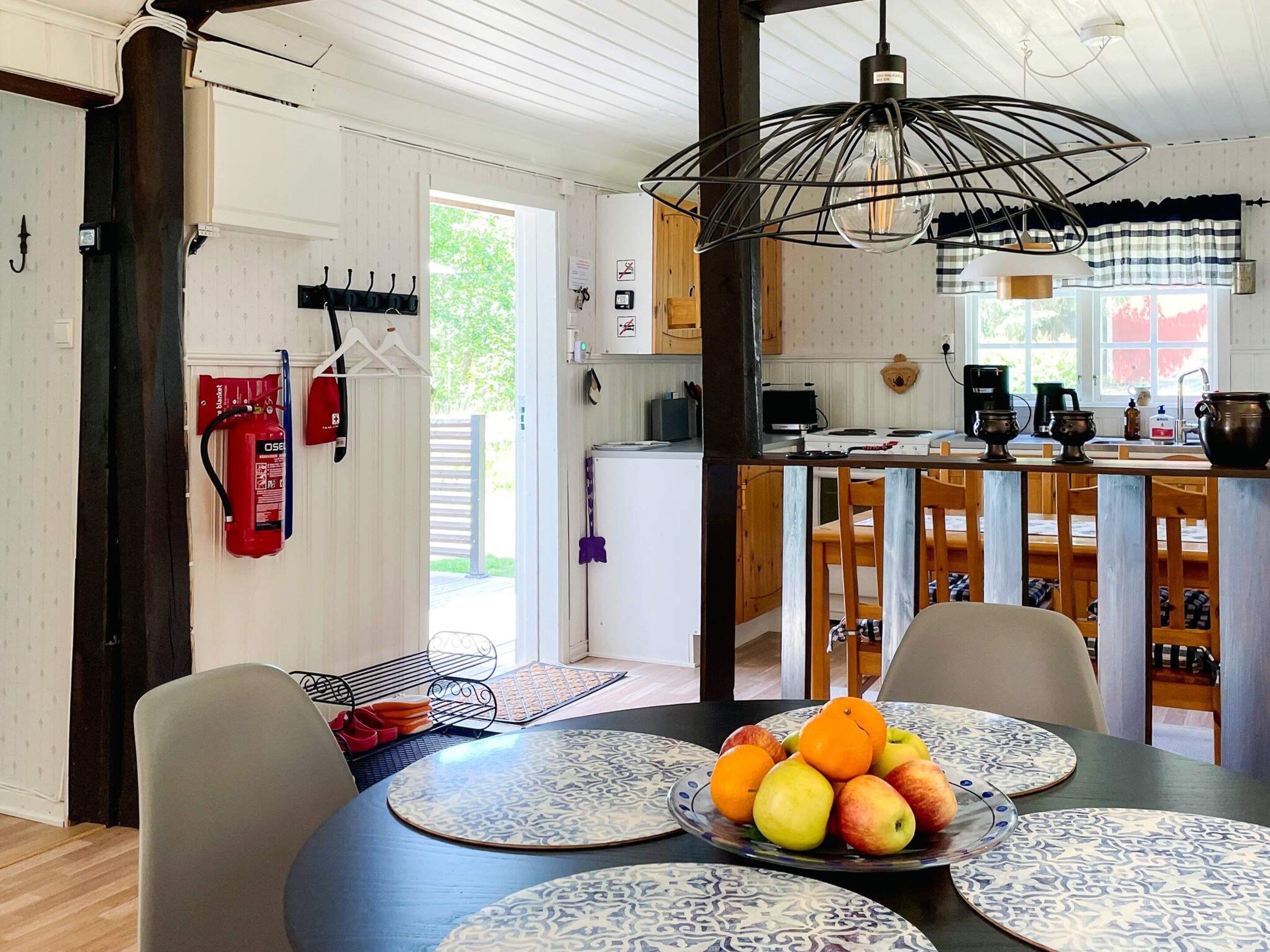 Un comedor con una mesa redonda dispuesta para cuatro personas, un frutero sobre la mesa y una cocina al fondo.