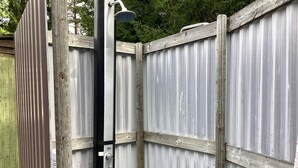 Una ducha exterior con estructura metálica y cabezal de ducha manual, adosada a una valla de madera.