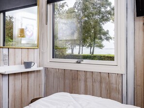 Une chambre à coucher avec un lit, une fenêtre donnant sur des arbres et de l’eau, une table de chevet avec une tasse, et une étagère murale avec un cadre photo.