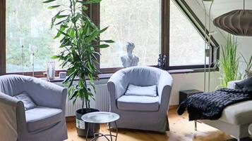 Una habitación con dos sillones grises, una mesita redonda, una planta en maceta y vistas a árboles a través de amplios ventanales.