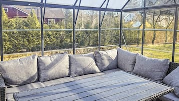 Un espacio de estar al aire libre techado con un banco y cojines, ventanas amplias y vistas a una casa y árboles.