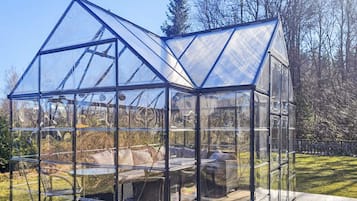 Estructura de invernadero transparente con techo y paredes, una banca en el interior y una terraza de madera en el exterior.