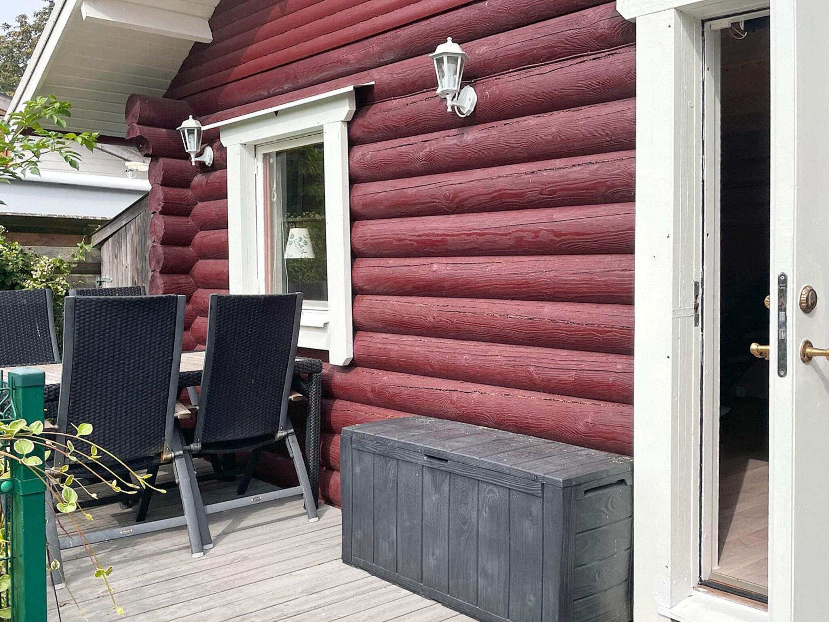 A log cabin with a patio area featuring two wicker tables and chairs, a small window with a white frame, and a door with a brass handle.
